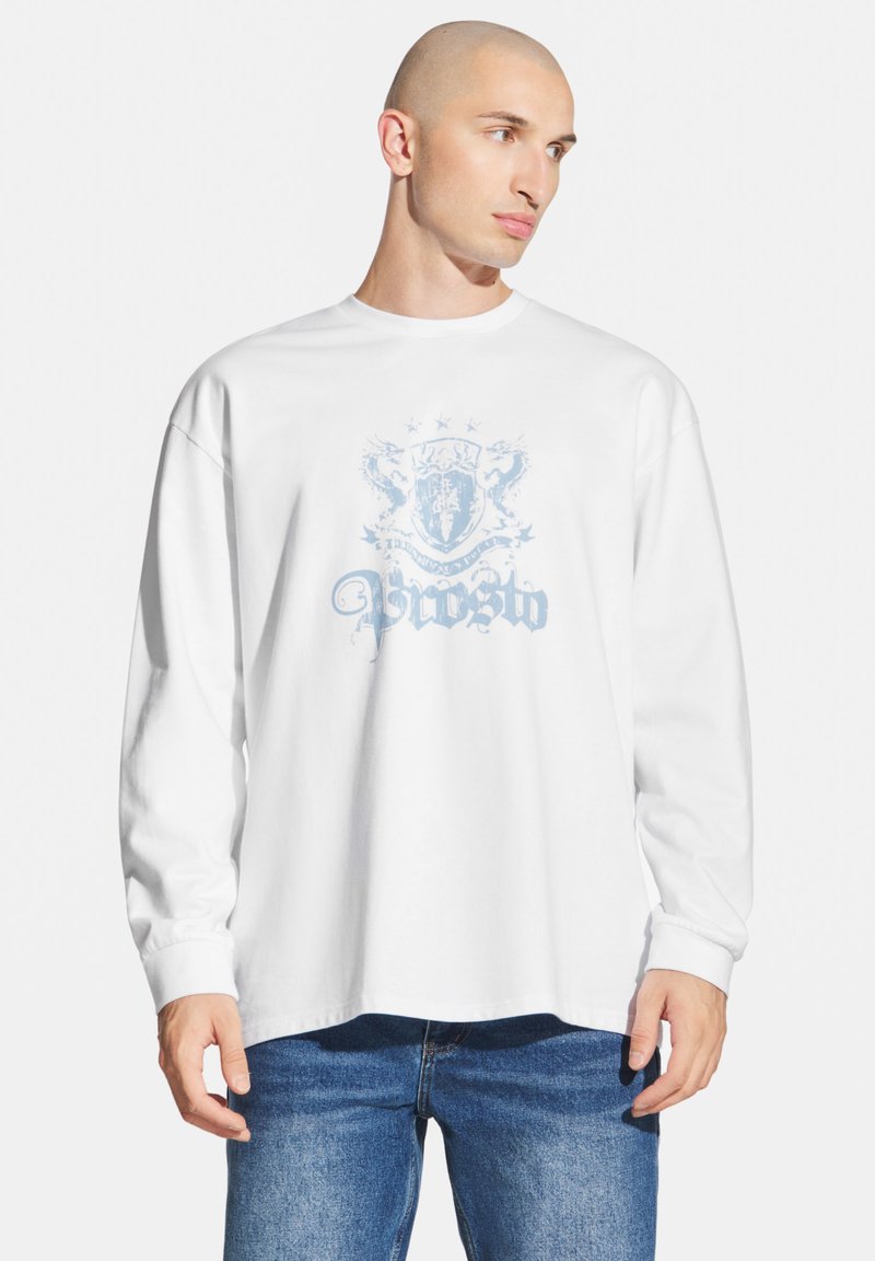 Biała koszulka z długim rękawem z jasnoniebieską grafiką przedstawiającą herb oraz ozdobny tekst z przodu. Luźny krój, miękki bawełniany materiał.