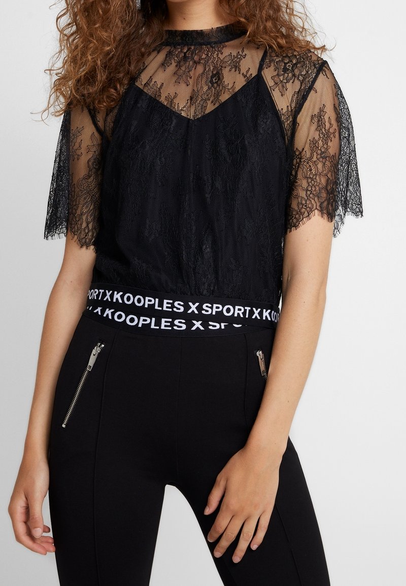 Femme portant un haut noir en dentelle à manches courtes avec un col et des manches transparentes, associé à un pantalon noir avec des poches zippées à l'avant.