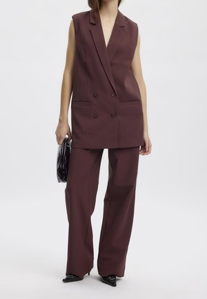 Kvinde iført ærmeløs, dobbeltradet bourgogneblazer og matchende bukser med vide ben, der holder en sort clutch og bærer sorte spidse sko.