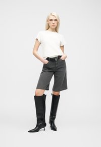 Jeune femme debout, mains dans les poches, portant un t-shirt blanc, un short gris au genou, une ceinture noire et des bottes noires à talons montant jusqu'aux genoux.