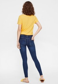 Kvinna med lockigt hår som bär en gul t-shirt, mörkblå slim fit-jeans och gula slip-on sandaler, stående med en hand i fickan.