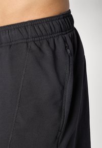 Shorts de sport noirs avec une taille élastique, une poche zippée sur le côté et une texture de tissu lisse. Présente des coutures flatlock pour plus de durabilité.
