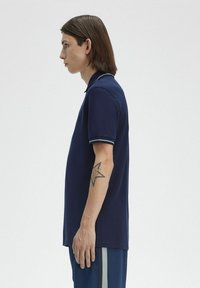 Polo de color navy con tela texturizada, mangas cortas y ribetes en azul claro que contrastan en el cuello y las mangas. Tatuaje notable en el brazo.