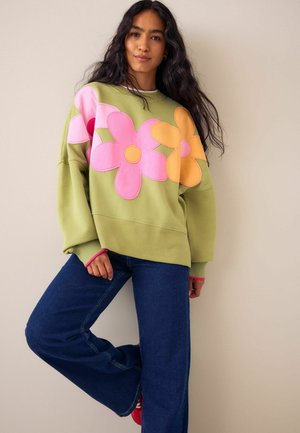 Kvinna i grön sweatshirt med stora rosa och orange blommönster, blå jeans, poserar med ett ben böjt mot en beige vägg.