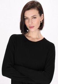 Zwarte longsleeve top met een ronde hals, gemaakt van zachte stof. Model toont minimalistische gouden oorbellen. Lichte achtergrond.