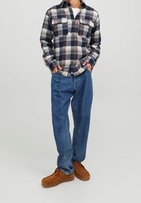 Camicia a quadri nei colori navy, crema e bordeaux con due tasche sul petto; abbinata a jeans blu larghi e scarpe in camoscio marroni.