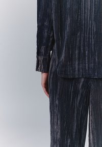 Sötét, texturált, passzoló szett függőleges csíkokkal, amely lazán illeszkedő ingből és bő szárú nadrágból áll. Az anyag puhanak és enyhén fényesnek tűnik.