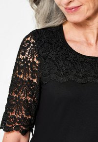 Zwarte top met een ingewikkelde zwarte kanten overlay op de korte mouwen, tekstuur met bloempatronen en een ronde halslijn.