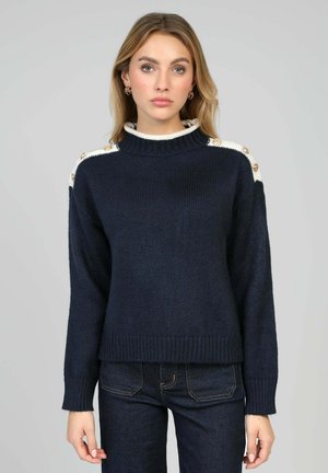 Pull en maille bleu marine avec un col montant bordé de crème, des boutons décoratifs aux épaules et des manches longues ; associé à un jean foncé.