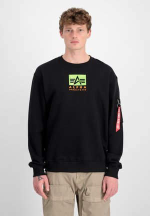 Zwarte sweatshirt met een groen en oranje logo, lange mouwen, geribbelde manchetten en een rode tag met tekst op de linker mouw.