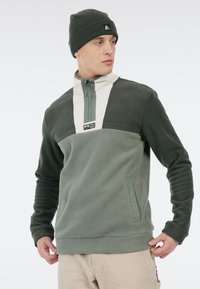 Fleece pullover in verschillende groentinten met een kwart-rits, een crèmekleurig borstpaneel en logo patch. Beschikt over zijzakken en een zachte textuur.