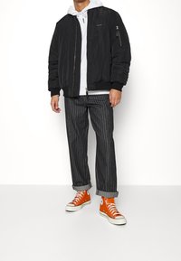 Černá bomber bunda s žebrovanými manžetami, bílá mikina s kapucí, pruhované šedé kalhoty a oranžové kotníkové tenisky. Na bundě je kapsa na zip.