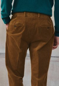 Pantaloni din catifea groasă maro, cu croi drept, dotati cu două buzunare spate cu închidere prin nasture și o textură netedă.