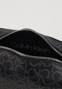 Černá kožená taška s texturou, která má stříbrný zip, s embosovaným logem Calvin Klein a opakujícím se vzorem CK na vnějším dojmu.