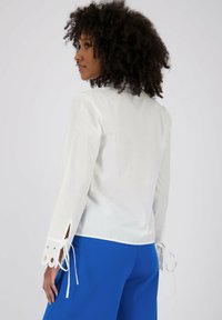 Camicia bianca a maniche lunghe con dettagli con tagli al polsino, abbinata a pantaloni blu. Presenta una vestibilità comoda e una texture liscia.