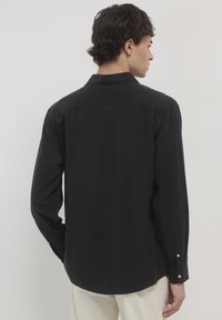 Mann mit dem Rücken zur Kamera, der ein langärmeliges schwarzes Hemd und cremefarbene Hose vor einem schlichten hellen Hintergrund trägt.