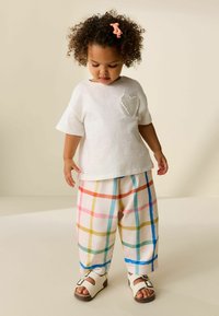 Weißes kurzärmeliges Hemd mit einem strukturierten Herzdetail, kombiniert mit bunten karrierten Hosen mit rosa-, blau- und gelben Streifen, getragen mit weißen Sandalen.