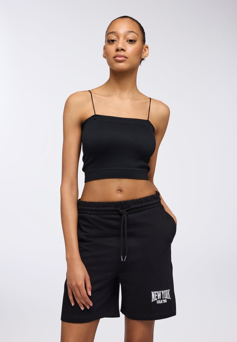 Top corto nero a coste con spalline sottili, abbinato a pantaloni corti neri con cordino che presentano un dettaglio stampato "NEW YORK USA 1996".
