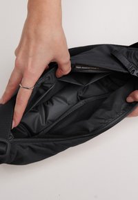Sorte nylon bæltetaske med en glat overflade. Lynlåsåbning afslører et quiltet indre. Har en justerbar strop til sikker bæring.