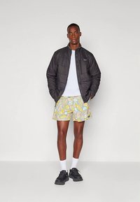 Άνδρας που στέκεται φορώντας ένα μαύρο τζάκετ με καπιτονέ σχέδιο, λευκό μπλουζάκι, λουλουδάτες κίτρινες και μπλε βερμούδες, λευκές κάλτσες και μαύρα αθλητικά παπούτσια σε λευκό φόντο.