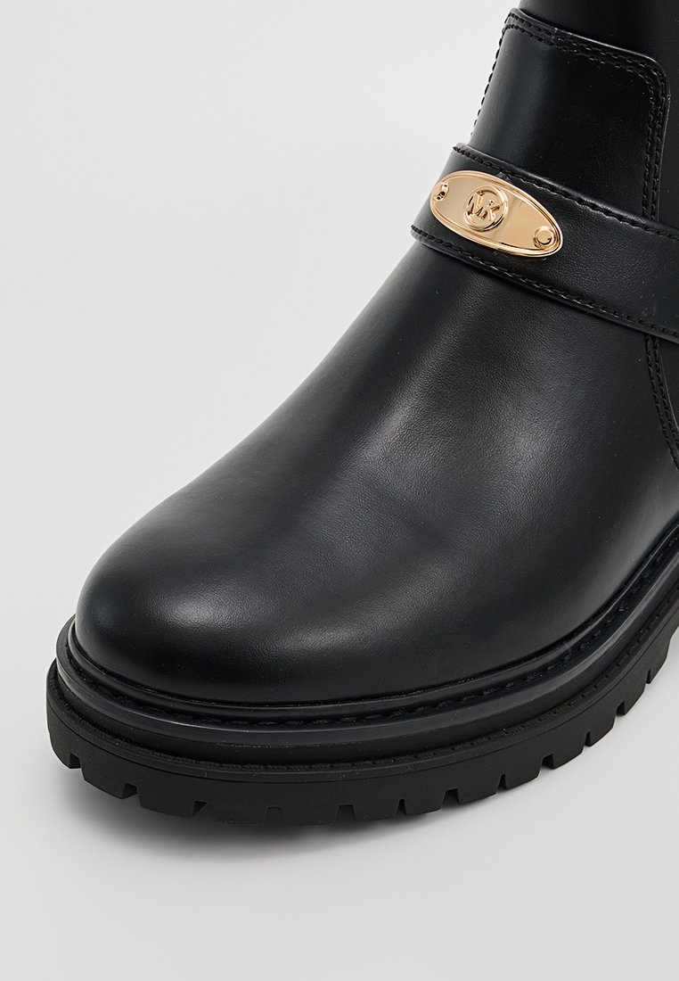 Czarne skórzane botki na kostkę z grubą podeszwą i złotą, owalną plakietką z logo na pasku powyżej palców.