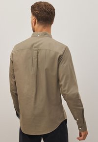 Homme portant une chemise beige à manches longues et à boutons, vu de dos sur un fond clair uni.