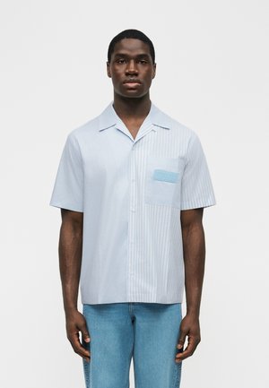 Male model wearing light blue and white short-sleeve button-up shirt with vertical stripes and patch pocket, paired with light blue jeans.