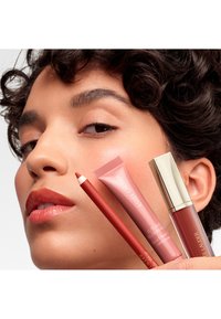 Czerwona konturówka do ust, różowy kremowy róż w tubce i brązowy błyszczyk do ust, prezentowane na jasnym tle, trzymane blisko twarzy modelki z kręconymi włosami.