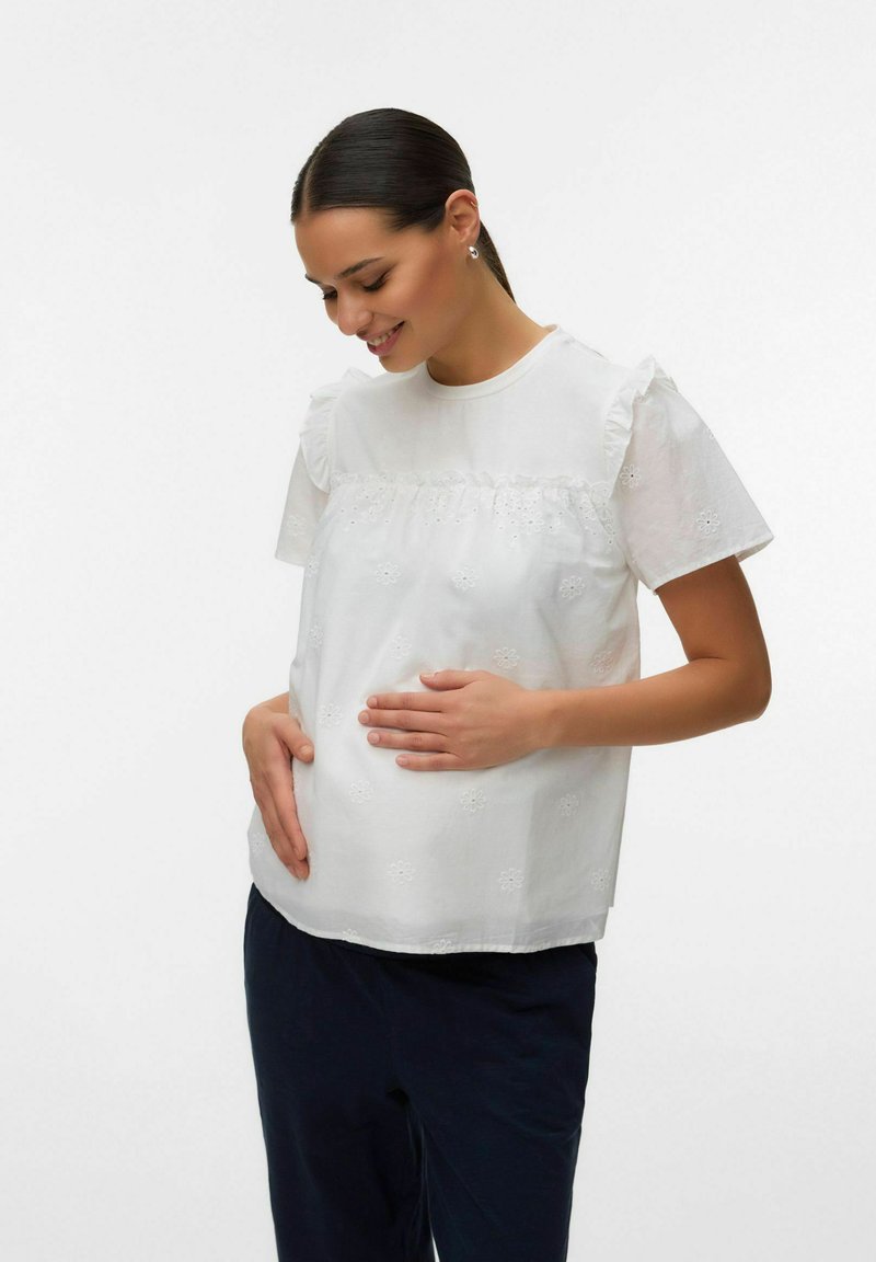 Femme enceinte aux cheveux foncés, portant un chemisier blanc brodé et un pantalon foncé, souriante et tenant son ventre avec ses deux mains.