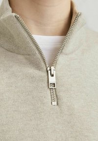 Svetar svetlo bež boje sa rajsferšlusom na zip, visokim ovratnikom, debelim rajsferšlusom i rebrastim detaljima duž ovratnika i donjeg dela. Mekani pleteni materijal.