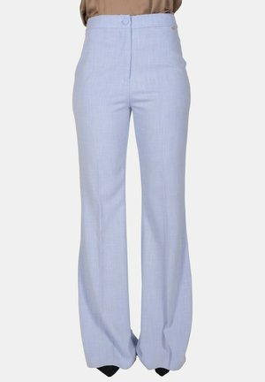 Persona con pantaloni a vita alta, azzurri e larghi, e una camicetta marrone infilata nei pantaloni, in piedi contro uno sfondo chiaro e semplice.