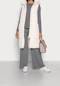 Elfenbensfärgad pufferväst med huva, randig långärmad topp, grå vida byxor och vita sneakers, håller en ljusrosa handväska.