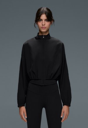 Donna con capelli scuri che indossa una giacca nera con zip e leggings neri, in piedi su uno sfondo grigio.