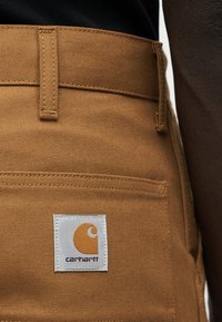 Κοντινό πλάνο καφέ παντελονιών Carhartt που δείχνει την πίσω τσέπη και το λογότυπο με το λευκό σήμα στην τσέπη.