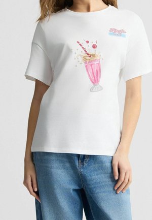 Camiseta blanca de manga corta con gráfico de batido rosa, dos cerezas y texto "Lucy Jo’s Diner", usada con vaqueros azules en una persona de pie.