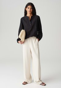 Camicia nera a maniche lunghe con una fila di bottoni, abbinata a pantaloni bianchi larghi e sandali neri. Tiene una borsa a tracolla beige con texture.