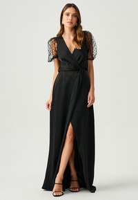 Black wrap dress with a deep V-neck, sheer puff sleeves featuring polka dots, and a thigh-high slit, made from a smooth fabric.