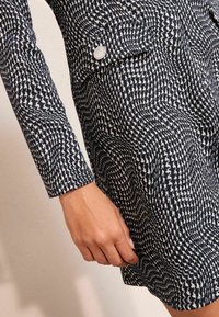 Rochie din țesătură cu model alb-negru, având un design geometric, cu un accent de nasture pe piept și un stil de mânecă ajustat.