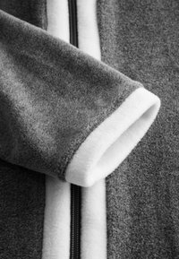 Graue Fleecejacke mit weißem Futter und Akzenten. Verfügt über einen glatten Reißverschluss in der Mitte und strukturierten Stoff an den Ärmeln.