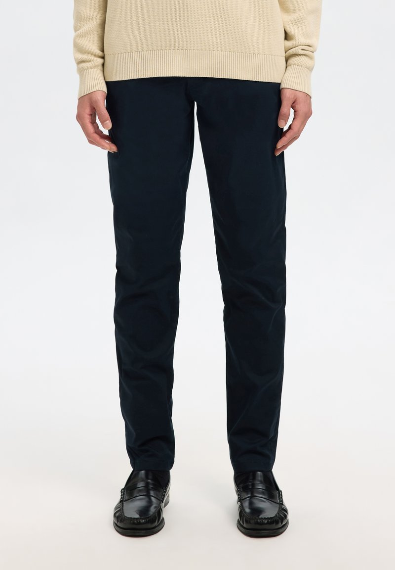 Person, die schwarze Hosen, schwarze Lederloafers und einen beigen Strickpullover trägt, steht vor einem schlichten hellen Hintergrund.