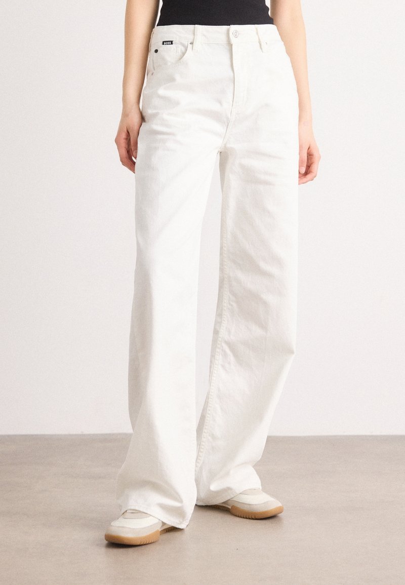 Femme vêtue d’un pantalon blanc taille haute, ample, avec un haut noir et des baskets blanches à semelles marron clair.