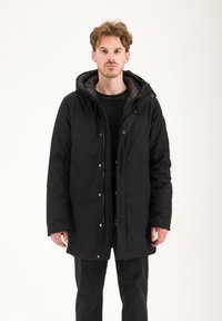 Parka nera isolata con chiusura a zip e bottoni a pressione, cappuccio regolabile e tasche laterali, indossata sopra un maglione scuro e pantaloni.