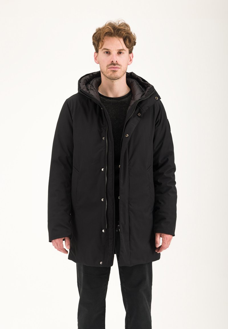Parka nera isolata con chiusura a zip e bottoni a pressione, cappuccio regolabile e tasche laterali, indossata sopra un maglione scuro e pantaloni.