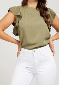 Olivengrøn bluse med flæser og korte ærmer, glat tekstur, parret med højtaljede hvide jeans med en guldknap og lommer.