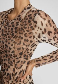 Nära bild av en person som bär en långärmad, brunt och svart leopardmönstrad mesh-top med puffärmar och synliga sömdetaljer.