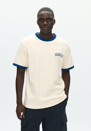Camiseta crema con cuello redondo y puños de manga en azul marino, con "DOUBLE V" bordado en azul oscuro en el pecho izquierdo.