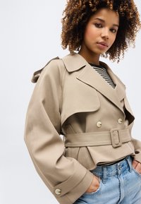 Νεαρή γυναίκα με σγουρά μαλλιά φοράει μπεζ καμπαρντίνα με ζώνη πάνω από μπλούζα με ρίγες και μπλε τζιν, ποζάροντας με τα χέρια στις τσέπες μπροστά σε ανοιχτό φόντο.