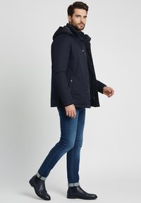 Hombre vistiendo una chaqueta con capucha azul marino, jeans ajustados azules y botas de cuero oscuras hasta el tobillo, caminando contra un fondo gris claro liso.