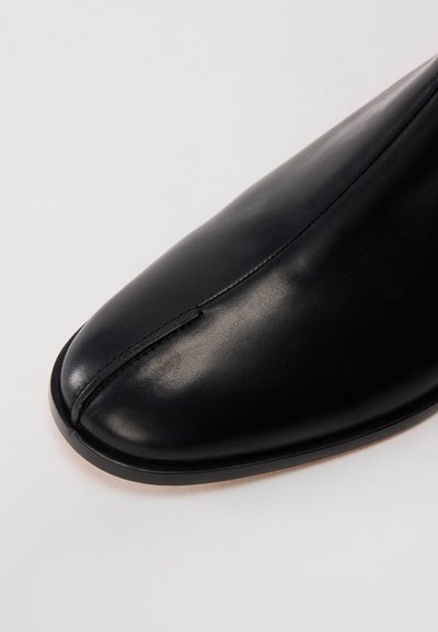 Nahaufnahme einer schwarzen Leder-Schuhspitze mit einer mittigen Naht und glatter Oberfläche auf einem hellen Hintergrund.