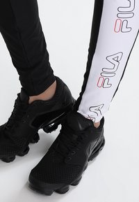 Person som bär svarta Nike-sneakers och svarta och vita Filaleggings, står på ett vitt golv, med fötterna nära varandra.